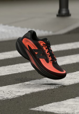 Must ja erkoranž New Balance jooksujalats õhus üle kõnniteel asfaldil, millel on valged triibud.