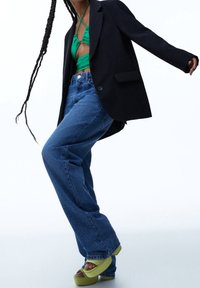 Svart kavaj över en grön cropped topp, kombinerad med högmidjade blå jeans och limegröna plattformssandaletter. Detaljerade texturer och färger finns närvarande.