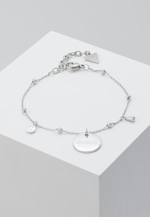 Delikat sølvkædearmbånd med små perler og tre vedhæng, inklusive et rundt vedhæng med indgraveret "GUESS" og en hummerlåse.