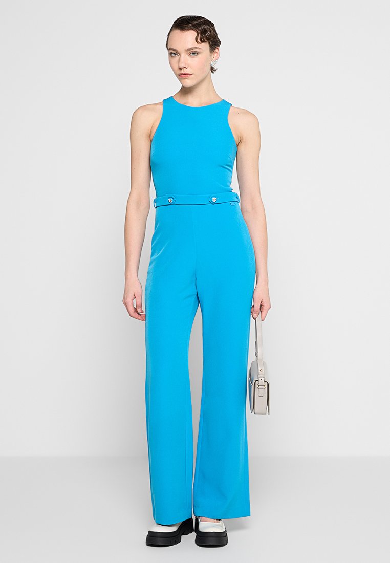 Vrouw in een felblauwe mouwloze jumpsuit met uitlopende pijpen, met een kleine grijze handtas, staand tegen een effen witte achtergrond.