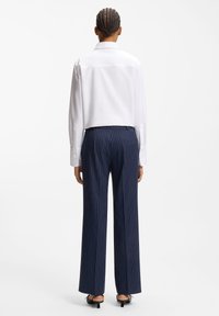 Chemise blanche à manches longues avec une pince dans le dos, associée à un pantalon bleu marine à fines rayures blanches, doté d'une coupe ajustée et de jambes évasées.