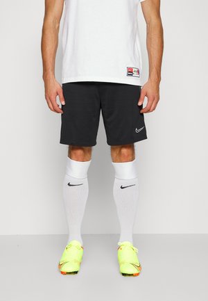 Homme portant un short noir Nike, des chaussettes blanches Nike montantes, des crampons de football jaune néon et un t-shirt de sport blanc avec une petite étiquette.