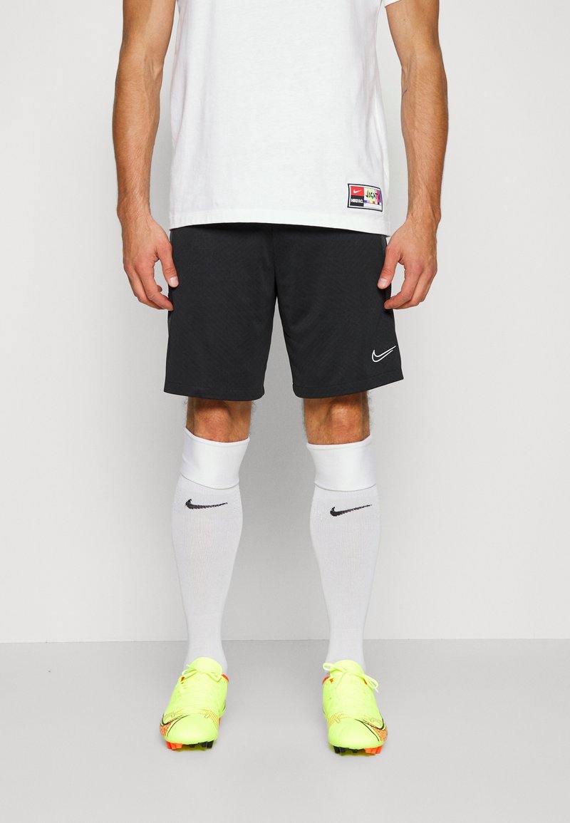 Homme portant un short noir Nike, des chaussettes blanches Nike montantes, des crampons de football jaune néon et un t-shirt de sport blanc avec une petite étiquette.