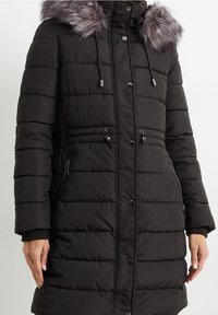 Svart puffjacka med fuskpäls på huven, horisontellt kviltat mönster, dragsko i midjan och fickor med blixtlås på sidorna. Har tryckknapp och blixtlåsförslutning.