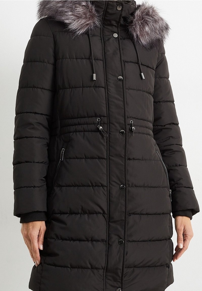 Svart puffjacka med fuskpäls på huven, horisontellt kviltat mönster, dragsko i midjan och fickor med blixtlås på sidorna. Har tryckknapp och blixtlåsförslutning.