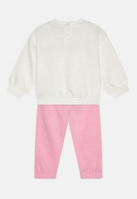 Sudadera blanca con puños acanalados, que presenta un cierre de botón en la parte posterior. Pantalones de chándal rosas con puños elásticos y textura suave.
