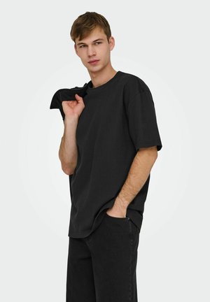 Młody mężczyzna w czarnej luźnej koszulce i spodniach, trzymający kurtkę na ramieniu, z jedną ręką w kieszeni, na białym tle.