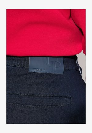 I jeans in denim blu scuro presentano una distintiva toppa in pelle sulla vita con testo in rilievo. Il tessuto ha una consistenza liscia.