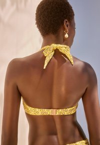 Sárga bikini felsőrész mintás dizájnnal, nyakba akasztható, hátul megkötős és fém kapoccsal záródik. Simára és textúrált anyagból készült.