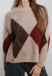Maglione rosa tenue con grandi motivi a rombi marroni e rossi indossato da una persona con lunghi capelli castani e pantaloni scuri.