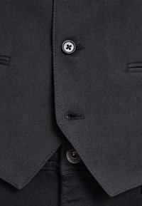 Tummanharmaasta kankaasta valmistettu liivi, jossa on teksturoitu kudos, pyöreä kaula-aukko, kaksi etutaskua ja mustat napit, joissa on neljä reikää.