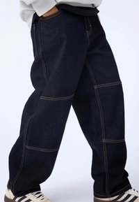 Dark blue denim cargo pants featuring visible stitching, loose fit, side pockets, and a slightly tapered leg. Designed for casual wear.