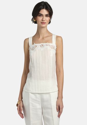 Donna che indossa una canotta bianca senza maniche con pieghe verticali e rifiniture in pizzo, abbinata a pantaloni bianchi e gioielli dorati, in piedi contro uno sfondo semplice.
