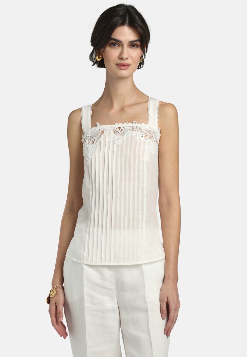 Femme portant un haut blanc sans manches avec des plis verticaux et une bordure en dentelle, associée à un pantalon blanc et des bijoux en or, debout devant un fond uni.