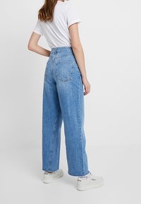 Světlé modré džínové džíny s vysokým pasem a volným střihem, s zadními kapsami a hladkou texturou, v kombinaci s bílými teniskami.