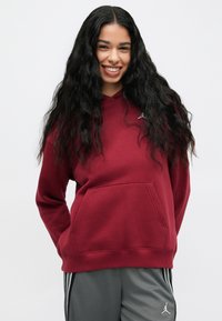 Smilende kvinne med langt, krøllete svart hår, iført en maroon hettegenser og grå sportsbukser med hvite striper mot en ensfarget bakgrunn.