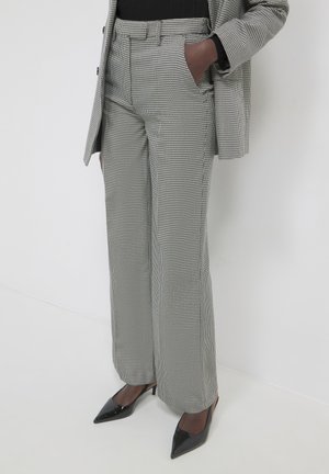 Persona che indossa pantaloni a gamba larga in tessuto tartan bianco e nero e un blazer abbinato, con tacchi a punta neri e una mano in tasca.