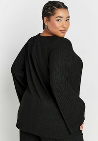 Musta teksturoitu paita, jossa on pitkät hihat ja rento istuvuus, hienovaraisella sahalaitakuviolla ja pyöreällä kaula-aukolla.