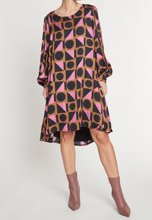 Frau, die ein locker sitzendes, knielanges Kleid mit geometrischem Muster in Schwarz, Braun und Pink trägt, kombiniert mit mauvefarbenen Stiefeletten, stehend vor weißem Hintergrund.