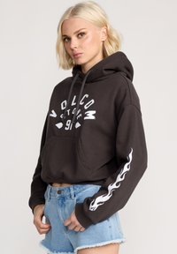 Haut à capuche court marron foncé avec logo Volcom blanc et motif de flammes le long de la manche gauche. Tissu doux et poche kangourou.