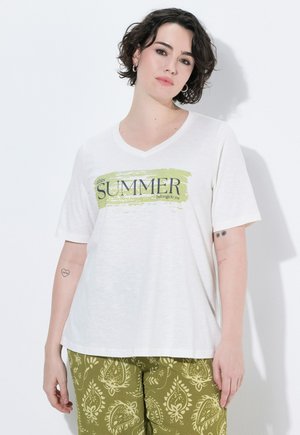 Person med kort mørkt hår iført en hvid t-shirt med teksten "this SUMMER belongs to you" og grønne mønstrede bukser, der står op ad en hvid væg.