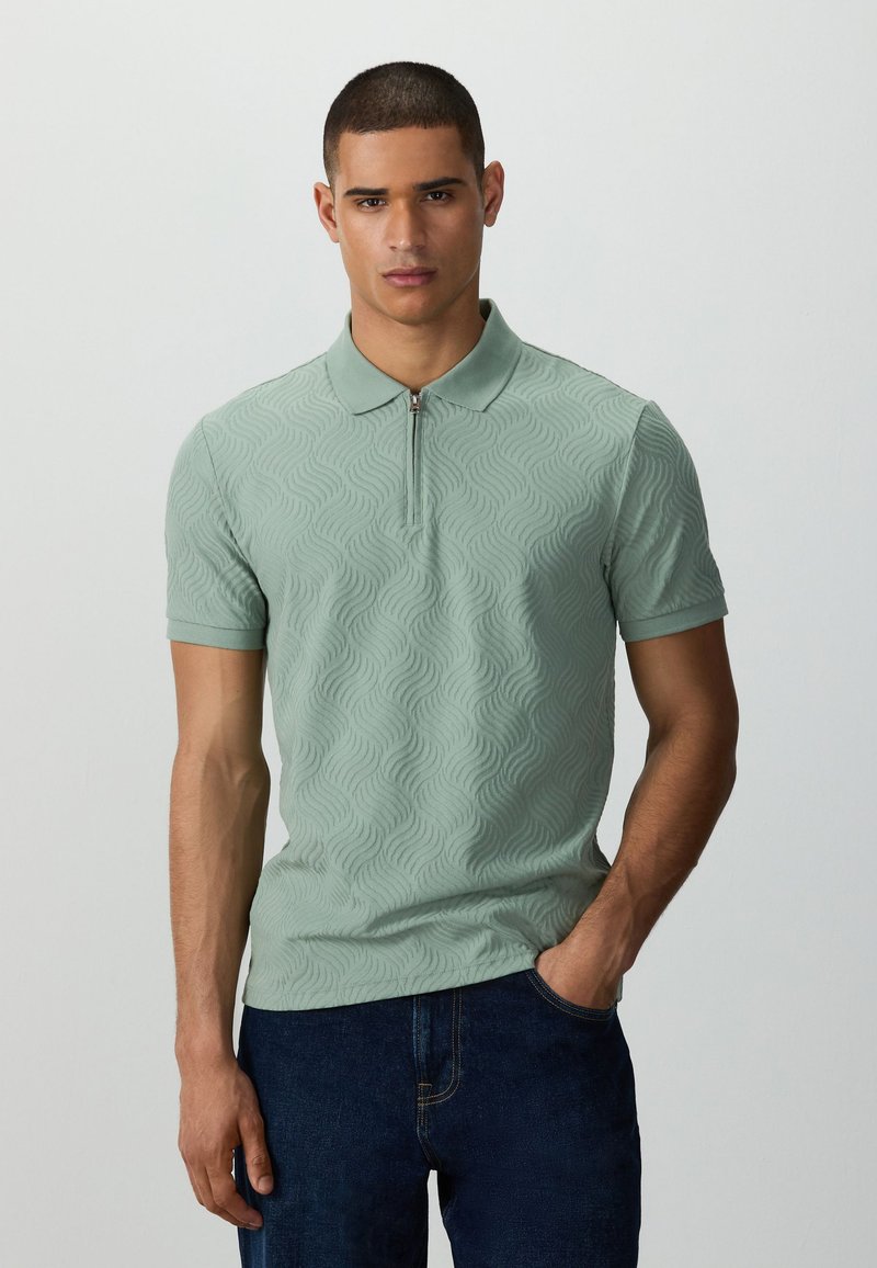Homme portant un polo vert clair à manches courtes avec un motif texturé ondulé et un jean bleu foncé, debout avec une main dans la poche.