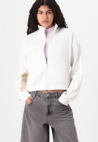 Hvid kort zip-up jakke lavet af blødt stof, med lange ærmer og et diskret logo på brystet, parret med grå rummelige jeans.
