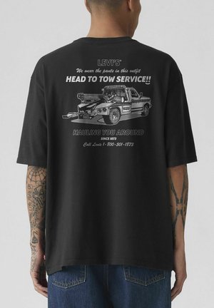 Person trägt ein schwarzes Levi's T-Shirt mit einer Abschleppwagen-Grafik und Text zum Head-to-Tow-Service auf der Rückseite, sichtbare Tätowierungen an den Armen.