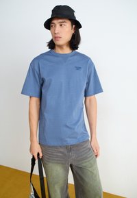 Tricou din bumbac albastru, cu mâneci scurte, decolteu rotund și un logo mic pe piept. Asortat cu blugi gri și o pălărie neagră de tip bucket.