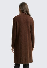 Kobieta o długich brązowych włosach, ubrana w długi, teksturowany brązowy kardigan, stoi tyłem do aparatu na jednolitym tle.