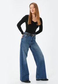 Schwarzes, geripptes Langarmoberteil, kombiniert mit hoch taillierten, weit geschnittenen blauen Jeans. Auffällige Details sind ein schwarzer Gürtel und elegantes Schuhwerk.