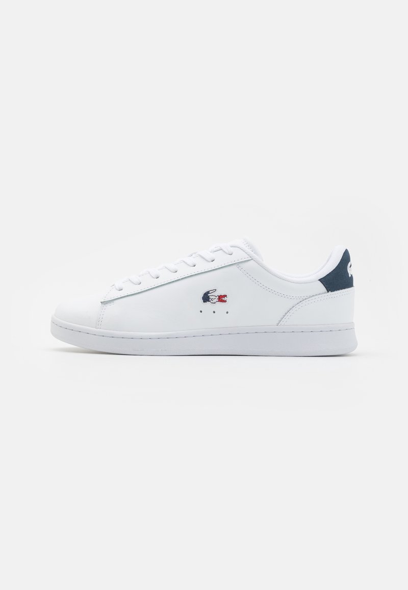 Baskets basses blanches avec lacets, languette de talon bleu marine et petit logo crocodile bleu et rouge sur le côté.