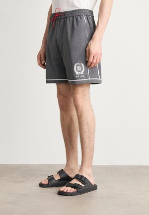 Homme portant un short gris à fines rayures avec un logo blanc et un cordon rouge, associé à des sandales noires à boucle sur un sol clair.