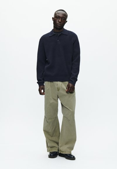 Mornarsko pleteno pulover z ovratnikom in tremi gumbi, v kombinaciji s širokimi, svetlo olivnimi cargo hlačami in črnimi gležnjarji.