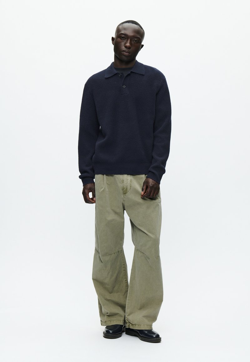 Maglione lavorato a maglia blu navy con colletto e tre bottoni, abbinato a pantaloni cargo larghi di un verde oliva chiaro e stivaletti neri fino alla caviglia.