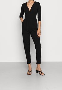 Woman wearing a black jumpsuit with a deep V-neck and three-quarter sleeves, paired with black strappy heeled sandals.