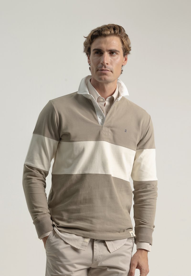 Camisa polo de manga larga a rayas en tonos beige y crema con un cuello alto, material de algodón suave y un pequeño logotipo en el pecho. Combinada con pantalones claros.