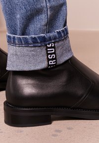 Svarta läderankelboots som bärs med uppvikta blå jeans som visar en "VERSUS"-etikett på kanten.