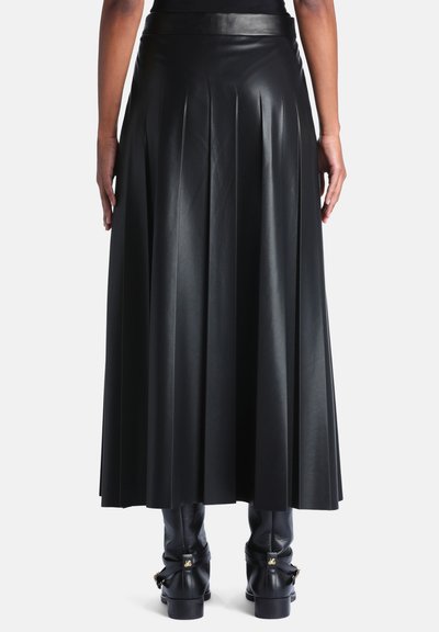 Personne portant une longue jupe noire plissée en cuir et des bottines noires, debout devant un fond blanc uni.