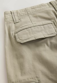 Nærbillede af beige cargo-bukser med baglomme med klap og bæltestrop på blødt stof.