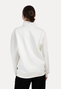Sudadera de forro polar blanco con alto cuello, puños acanalados y dobladillo. Presenta una etiqueta lateral y un corte holgado. Textura lisa, sin patrones visibles.