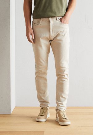 Lyse beige denim jeans med slim fit, der har fem lommer og diskrete syninger, kombineret med beige sneakers og en grøn skjorte.