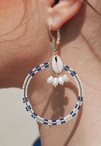 De grandes boucles d'oreilles en forme de créole avec des perles blanches, des accents bleus et des garnitures dorées. Un centre en forme de coquillage avec des pendentifs en perles ajoute de l'élégance.