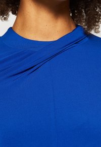 Blå topp med ett samlat detalj vid halsringningen, som har en mjuk och elastisk textil samt en figurnära design. Närbild av tygtexturen.