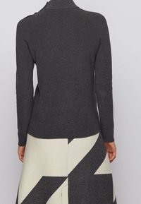 Pull côtelé gris foncé avec un col montant et des détails à boutons. Associé à une jupe crème ornée de motifs géométriques noirs.