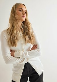 Vit knäpp cardigan med lång ärm, ribbade muddar och pärlemor knappar. Mjuk textur och lagerdesign med en lätt fall.