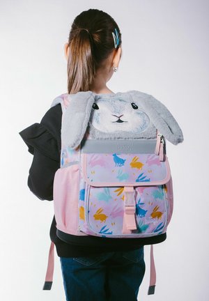 Ragazza con capelli castani raccolti in una coda di cavallo che indossa uno zaino rosa pastello con la faccia di un coniglio con orecchie cadenti e motivi colorati di conigli.