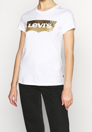 Osoba nosząca białą koszulkę z krótkim rękawem z logo Levi's w złotym brokacie, połączoną z czarnymi spodniami, na jasnym, jednolitym tle.