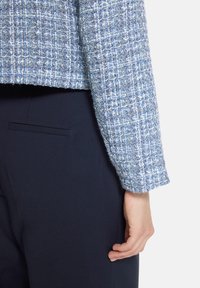 Femme portant une veste en tweed texturée bleu et blanc avec un pantalon ajusté bleu marine, main posée de manière décontractée sur le côté.