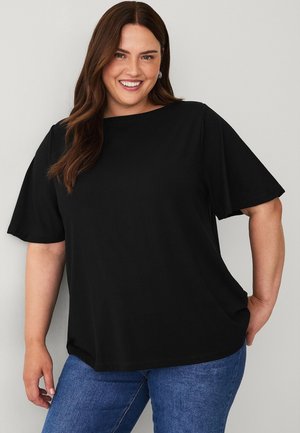 Smilende kvinde iført en løs sort kortærmet top og blå jeans, stående mod en lysegrå baggrund.